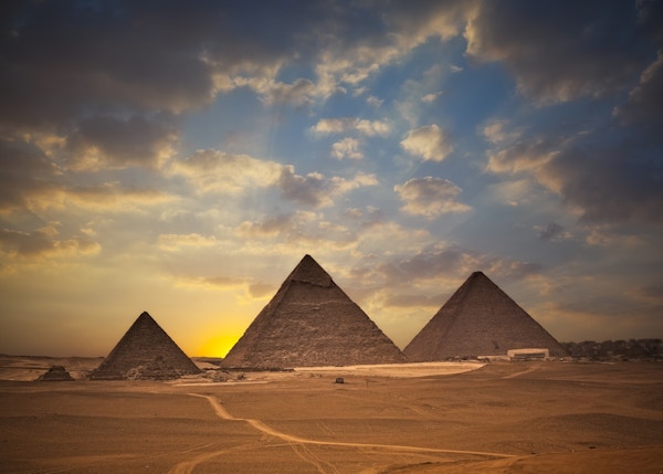Pyramidene i Egype fotografert på avstand med en stigende sol bak og lettskyet himmel.