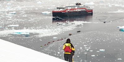 Skip som ligger ute blant is der småbåter og kajakker er på vei inn til land. Person står på land og ser utover med ryggen til.