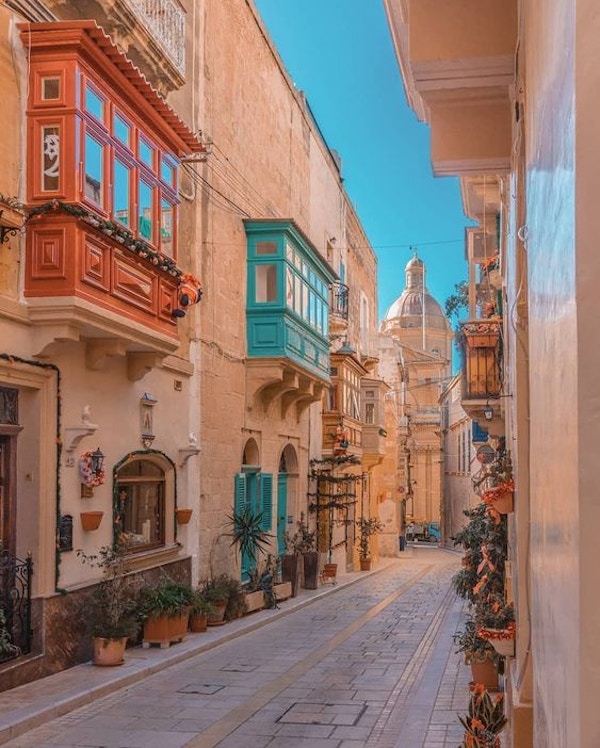 En gate i Birgu, Malta, med typiske maltesiske balkonger malt i flotte farger og domen på kirken i enden av gaten