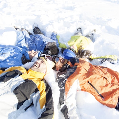 Ungdommer som ligger på snøen, holder hender og lager snøengler. De er ikledd skiklær, hansker og skibriller. Visning i høy vinkel.
