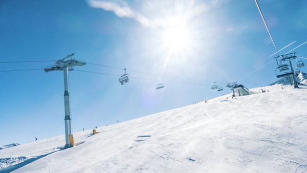 Folk beveger seg opp og ned i skråning med stolheis i skianlegg. Lav vinkel. Sollys og klar himmel på bakgrunn.