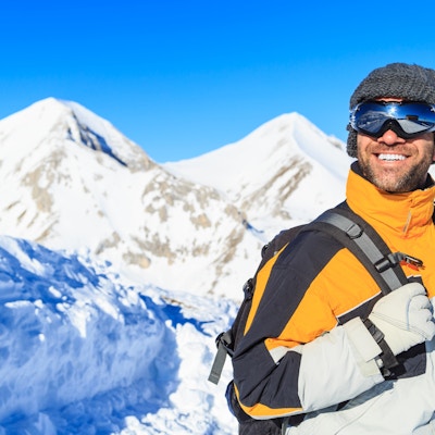 Munter ung mann som vandrer på snøfjellet. Med skidress, vernebriller, hansker og strikkelue, ryggsekk. I bakgrunnen en fjelltopp med snø.