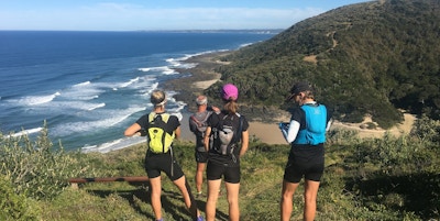 Fire vandrere står med ryggsekk og ryggen til på en høyde og ser utover havet
