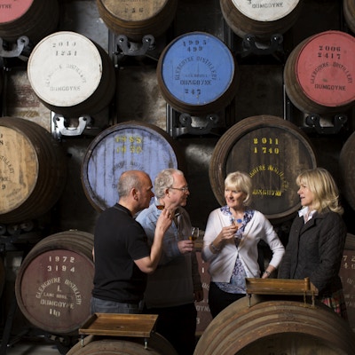 To menn og to kvinner er på whiskysmaking inne i et destilleri med tønner i bakgrunnen