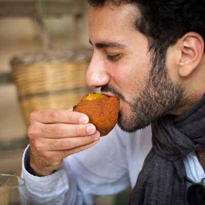 Sicilia: En ung siciliansk mann som biter i en tradisjonell siciliansk frityrstekt riskrokett (arancina). Nærbilde.