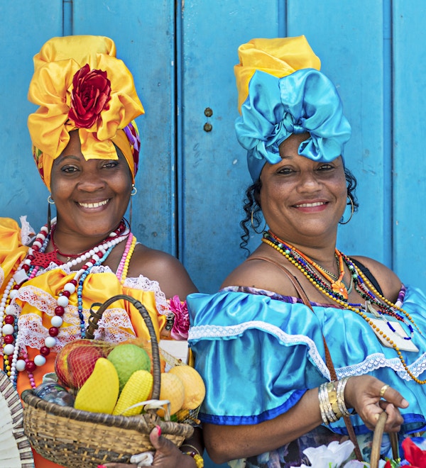 Portrett av glade cubanske kvinner som står mot blå trevegg. Smilende modne kvinner er i tradisjonelle kjoler. De er med fruktkurv og håndvifte.
