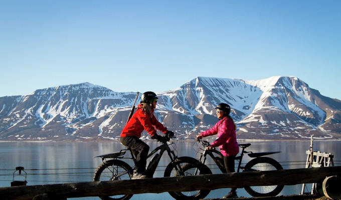 To jenter på sykler med tjukke hjul står med en fot i bakken og snakker. De har hjelm på og den ene har rifle