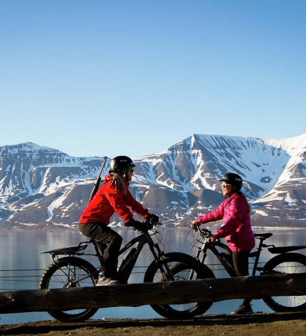 To jenter på sykler med tjukke hjul står med en fot i bakken og snakker. De har hjelm på og den ene har rifle