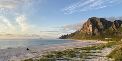 To mennesker sitterpå stol på sanden på stranden Bleik på Andøya og ser ut mot havet med fjell i bakgrunnen