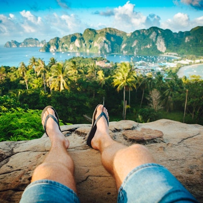 Mann sitter på fjellknaus og ser mot palmer, strand og hav
