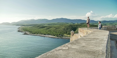 To damer står og ser utover utsikten over havet og Sierra Nevada-fjellene fra festningen El Morro ved innseglingen til Santiago de Cuba
